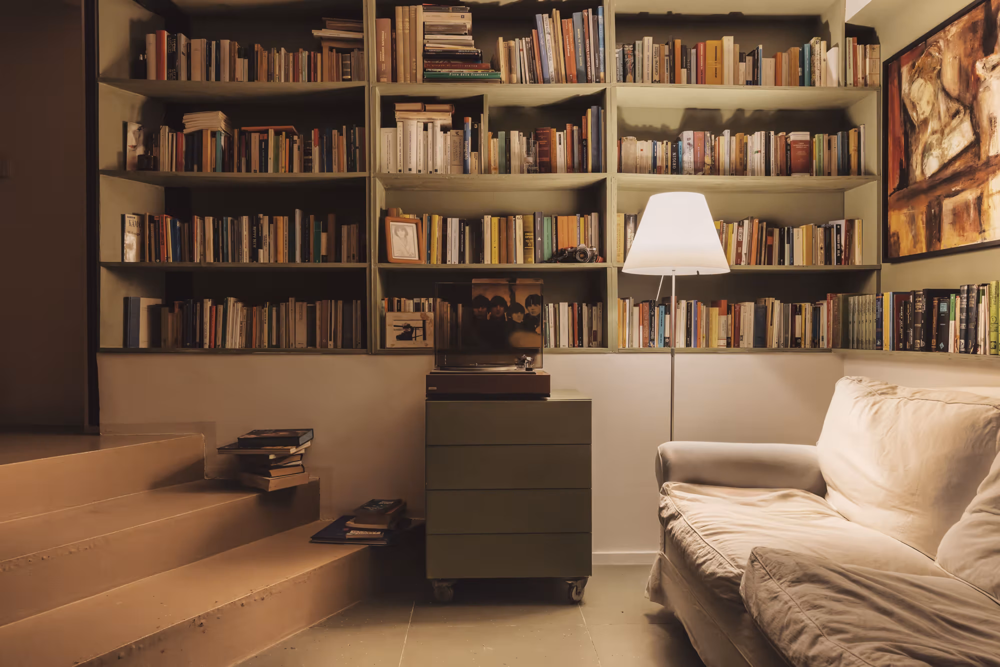 Sala comune con una libreria a tutta parete, un divano chiaro e una lampada da terra accesa