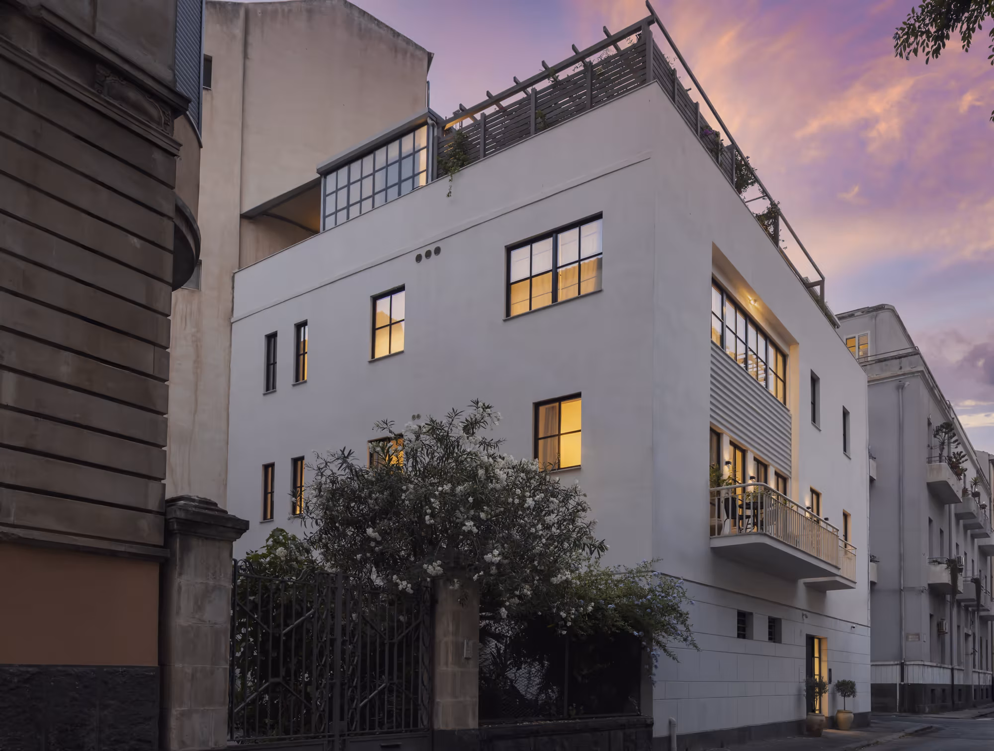 La facciata bianca della casa al tramonto, con le finestre illuminate e il giardino davanti